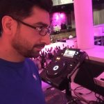 DJ Gilano steht vor einem DJ-Pult in einem legeren blauen T-Shirt
