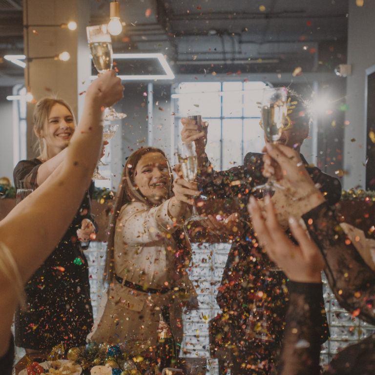 Vier Personen stoßen mit Sektgläsern. Goldender Konfetti fliegt in der Luft herum.