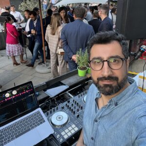 Mann im Jeanshemd lächelt in die Kamera. Im Hintergrund DJ-Pult und Menschen